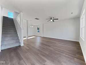A spacious, empty living room with light-colored walls, wood flooring, a ceiling fan, multiple windows, and a carpeted staircase on the left side. Natural light enters the room from the windows.