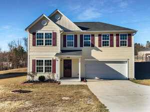 Two-story beige house with red shutters, a black front door, white trim, and a double garage. The driveway is concrete, and the yard is mostly grass with small shrubs and minimal landscaping. Trees are in the background.