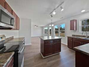 A modern kitchen with dark wood cabinets, stainless steel appliances, a central island, and adjoining dining area. Large windows provide natural light and offer views of an open grassy yard.