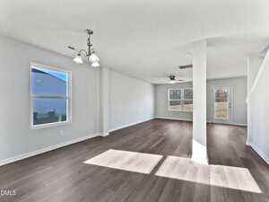 A spacious, empty room with light-colored walls, wood flooring, multiple windows letting in natural light, a ceiling fan, and a staircase to the right. The room has a clean, modern appearance.