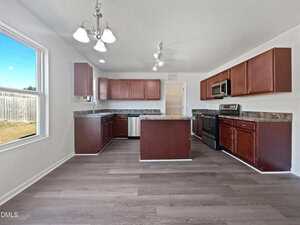 A modern kitchen with dark wood cabinets, stainless steel appliances, an island in the center, overhead lighting, and large window providing natural light. The floor is a light wood laminate.