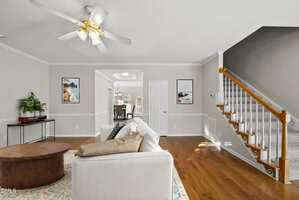 A living room with a white sofa, round wooden coffee table, two framed wall art pieces, hardwood floors, a staircase with a white railing, and a ceiling fan. Dining area visible through an open doorway.