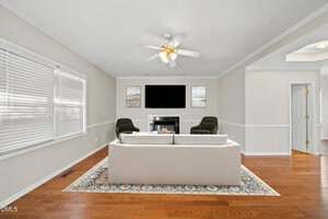 A living room with a white couch on a patterned rug, two green armchairs, a fireplace below a wall-mounted TV, and artwork above the fireplace. The room has hardwood floors, light walls, and a ceiling fan with lights.