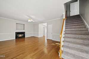 A spacious living room with light beige walls, wood flooring, a ceiling fan, a corner fireplace, and a staircase with carpeted steps and a wooden handrail leading to an upper floor.
