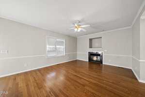 Empty living room with light beige walls, white trim, medium-tone hardwood floor, a ceiling fan with lights, two windows with blinds, and a fireplace set into a wall niche.