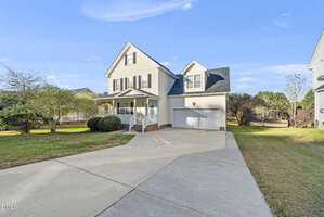 Two-story beige house with a covered front porch, attached two-car garage, and concrete driveway. The yard is grassy with bushes near the house, and trees are visible in the background under a blue sky.