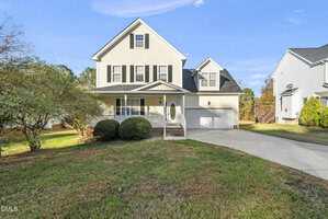 This two-story beige house at 1589 Rogers Pointe Lane in Creedmoor features black shutters, a covered front porch, an attached two-car garage, a grassy lawn, a driveway, and a small tree on the left side.
