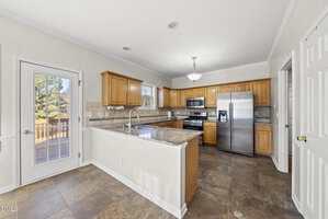 A spacious kitchen with wooden cabinets, stainless steel appliances, marble countertops, a double-door refrigerator, a central island with a sink, tile flooring, and a door leading to an outdoor deck.