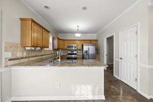 A modern kitchen with wooden cabinets, stainless steel appliances, granite countertops, tile backsplash, and a breakfast bar. A double sink is installed in the counter, and there is a pantry door on the right.