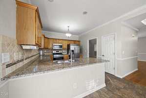 A kitchen with wooden cabinets, stainless steel appliances, granite countertops, a double sink, tiled floor, and a partial view of an adjacent room with hardwood floors.