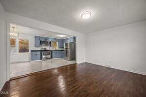 A spacious room with dark wood floors opens into a kitchen featuring blue cabinets, stainless steel appliances, tile flooring, and a window by the dining area. The walls are white, and ceiling lights are on in both areas.