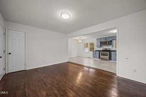 A view of an empty room with dark wood floors leading into a kitchen area. The kitchen features gray cabinets, stainless steel appliances, tile flooring, and a hanging light fixture over a dining space. White walls throughout.