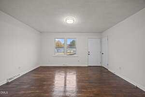 An empty room with white walls, dark wood flooring, a ceiling light fixture, two windows, and a white door. The room appears clean and well-lit with natural and artificial light.