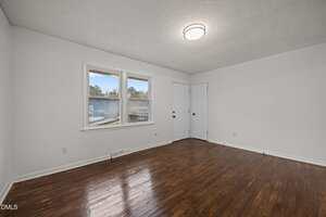 An empty room with white walls, a ceiling-mounted light fixture, two windows, two closed white doors, and dark hardwood floors. The room appears clean and well-lit.