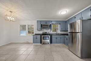 A kitchen with blue cabinets, stainless steel appliances, and a tiled floor. There is a window above the sink and a dining area with a light fixture on the left, next to two large windows facing outside.