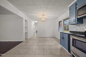 A kitchen with light beige tile flooring, blue lower cabinets, stainless steel appliances, white upper cabinets, and a modern light fixture. Large windows look out onto a yard; a hallway leads to other rooms.