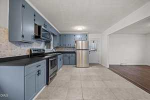 A kitchen with blue cabinets, stainless steel appliances, a tile backsplash, and beige floor tiles. The space opens into a room with wood flooring and white walls. A door with a window is visible in the back.