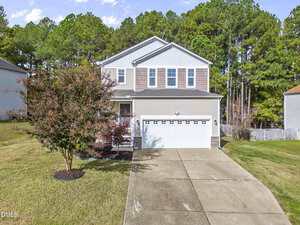 A two-story suburban house with beige siding and a white two-car garage sits on a sloped lawn at 1193 Shining Water Lane in Creedmoor, surrounded by tall pine trees and featuring a tree near the driveway.