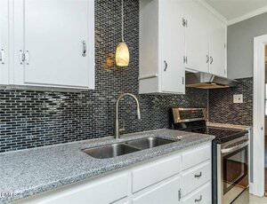 A modern kitchen with white cabinets, a gray granite countertop, a stainless steel sink, a silver electric stove, and a black and white mosaic tile backsplash. A pendant light hangs above the sink.