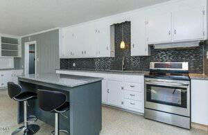Modern kitchen with white cabinets, stainless steel oven, and black tile backsplash. A gray island with a granite countertop has two black barstools. Upper and lower cabinets provide ample storage.