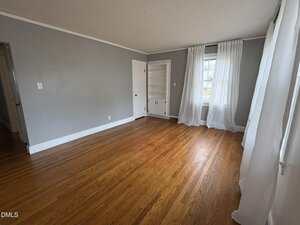 Empty room with gray walls, hardwood floors, a window with white curtains, a small built-in shelf, and two closed white doors. Natural light enters through the window on the right.