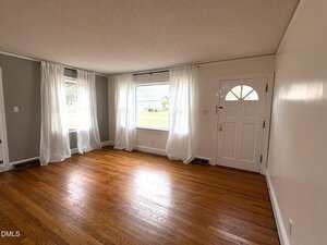 Empty living room with hardwood floors, white walls, two large windows with sheer white curtains, and a white front door with a window. Natural light fills the space, and the outside lawn is visible through the windows.
