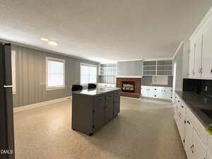 A spacious kitchen with gray paneled walls, white cabinets, a gray island with two black chairs, and a brick fireplace with built-in shelving. Large windows let in natural light.