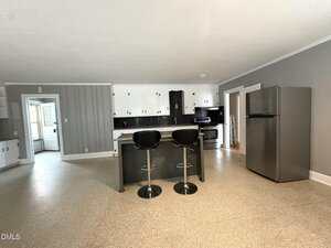 A modern kitchen with gray walls, white upper cabinets, black lower cabinets, a large island with two black barstools, stainless steel appliances, and beige flooring. A door leads to another room on the left.