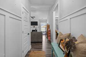 A view down a hallway with white walls and wood flooring, leading to a living room with a sofa, wall-mounted TV, and fireplace. A glass table with pillows and decorative branches is on the right.