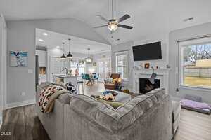 A modern living room with a large gray sectional sofa, a ceiling fan, a wall-mounted TV above a white fireplace, and adjacent open kitchen and dining areas. Large windows let in natural light.