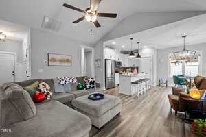 A modern living room with a gray sectional sofa, holiday decorations, open kitchen with an island and barstools, stainless steel appliances, and pendant lights, featuring wood flooring and neutral walls.