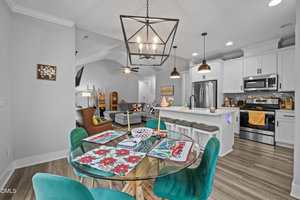 A modern open-concept kitchen and living area with a glass dining table, teal chairs, floral placemats, a white island with stools, stainless steel appliances, and neutral-toned furnishings and decor.