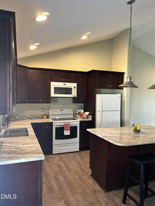 A kitchen with dark wood cabinets, white appliances including a stove, microwave, and refrigerator, marble-look countertops, an island with stools, and recessed lighting. A bowl of lemons sits on the island.