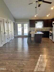 Open kitchen with dark cabinets, island with two stools, stainless steel appliances, ceiling fan, and double doors leading outside; wood flooring and natural light from windows and doors.