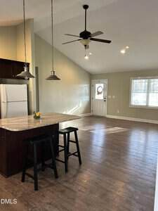 Open-concept kitchen and living area with light wood flooring, vaulted ceiling, ceiling fan, two pendant lights over a kitchen island with stools, and a white refrigerator; large window and door in the background.