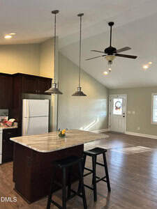 A kitchen with dark cabinets, a white refrigerator, and a marble island with two black stools. Pendant lights hang above the island, and a ceiling fan is on the vaulted ceiling in an open, empty living area.