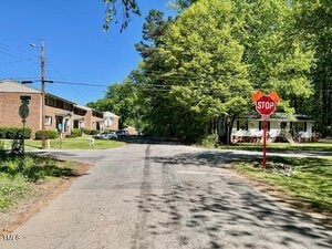 A quiet suburban street with apartment buildings on the left, a single-story house on the right, and a stop sign at the intersection. Trees line both sides of the road under a clear blue sky.