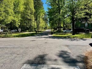 A residential street intersection is surrounded by trees with green foliage. There are houses and parked cars on both sides, and the scene is lit by sunlight. A stop sign is partially visible on the left.