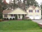 Located at 804 E E Street in Butner, this two-story yellow house with white trim features a covered front porch, brown shutters, a double garage, and is surrounded by neatly trimmed bushes on a spacious green lawn with scenic trees.