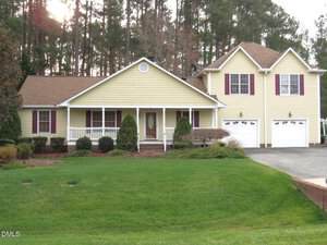 Located at 804 E E Street in Butner, this two-story yellow house with white trim features a covered front porch, brown shutters, a double garage, and is surrounded by neatly trimmed bushes on a spacious green lawn with scenic trees.