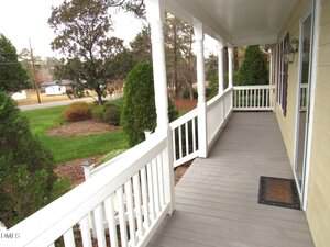 A covered front porch with white railings and columns overlooks a neatly maintained lawn with shrubs and trees. The porch has gray flooring and leads to the front door of a light yellow house.