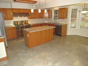 A spacious kitchen with wooden cabinets, stainless steel appliances, a large center island, tile flooring, pendant lights above the island, and a bay window area with natural light.