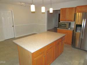 A kitchen with a center island, wooden cabinets, stainless steel refrigerator, countertop toaster oven, tiled floor, and three pendant lights hanging from the ceiling. A door and an open doorway are visible in the background.