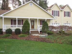 A yellow house with white trim features a covered front porch, brick steps, maroon shutters, a wooden front door with glass, attached two-car garage, and landscaped shrubs and trees in the front yard.