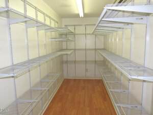 A large walk-in pantry with white wire shelving on both sides and at the end, light-colored walls, and a wood-look floor. A fluorescent light fixture is mounted on the ceiling.