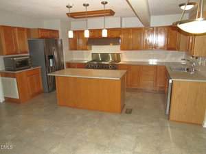 Spacious kitchen with tile flooring, wooden cabinets, stainless steel appliances, a large center island, double sink, and pendant lighting over the island.