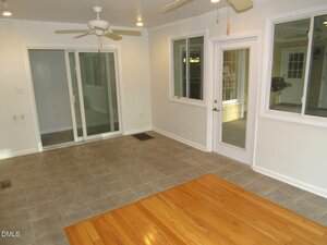 A room with light gray tile flooring transitioning to light wood flooring, white walls, a ceiling fan, sliding glass doors, a window, and a white door with a glass panel. The room is empty and well-lit.
