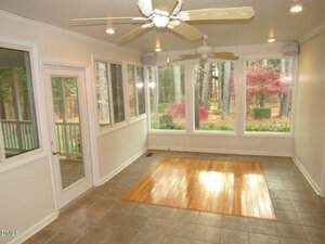 A sunroom with large windows on three sides, a tiled floor with a central section of wood flooring, two ceiling fans, and a view of trees and shrubs outside.