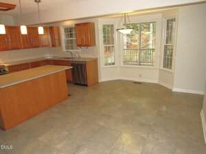 A kitchen with wood cabinets, stainless steel appliances, and an island opens to a tiled dining area with bay windows overlooking a wooded backyard. Pendant and ceiling lights are visible.