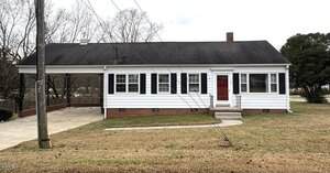 A single-story white house with black shutters, a red front door, and a brick foundation sits at 107 Grace Street, Oxford. The property features a driveway with a carport on the left, grassy front yard, and utility wires crossing in front.